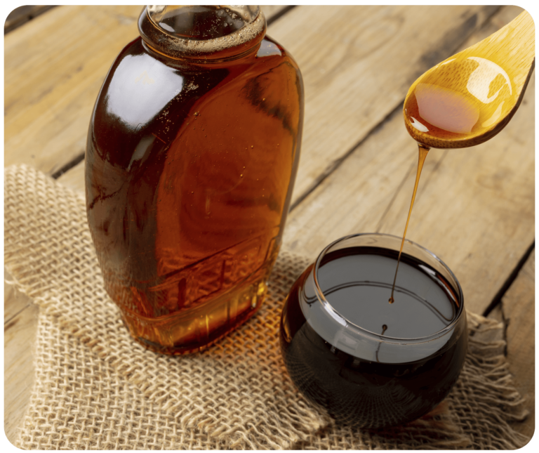 A bottle of maple syrup placed on a wooden table, adjacent to a glass bowl from which a ladle is scooping up the golden syrup, highlighting the rich, amber color of the maple syrup against the rustic backdrop.